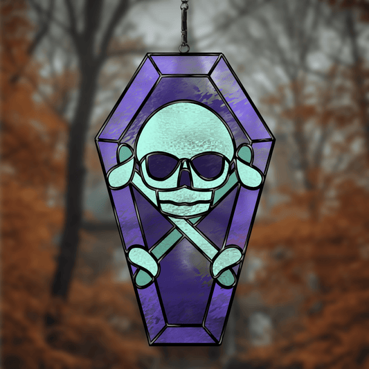 Stained glass window hanging with a skull design in a coffin, set against a blurred autumn forest background.